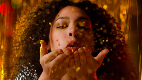 Young-Woman-Celebrating-At-Party-Or-Club-Blowing-Handful-Of-Gold-Glitter-Towards-Camera